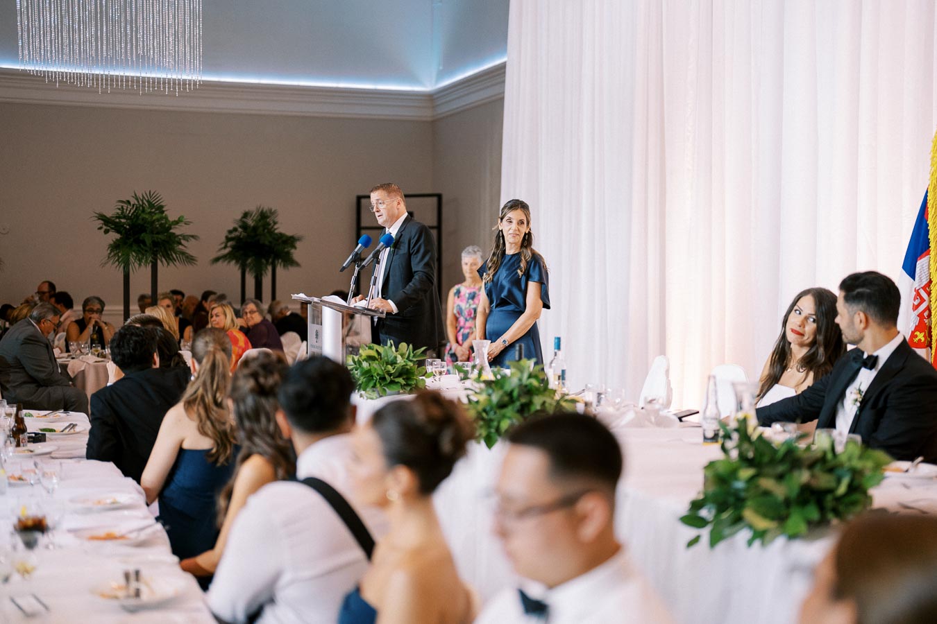 A man giving a speech at a formal event, standing at a podium with microphones in a decorated banquet hall, surrounded by elegantly dressed guests seated at long tables with greenery centerpieces.