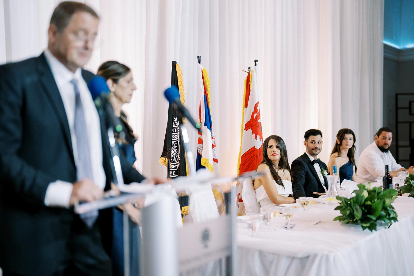 Group of people seated at a formal event table with national flags in the background, focused on a speaker in a suit at a podium, indoor setting.