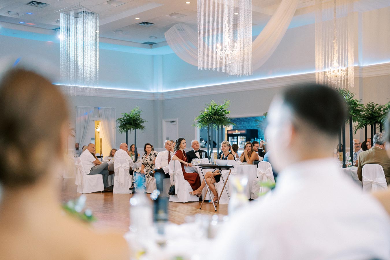 Elegant wedding reception with guests seated in a beautifully decorated ballroom, featuring chandeliers, white drapery, and greenery centerpieces.