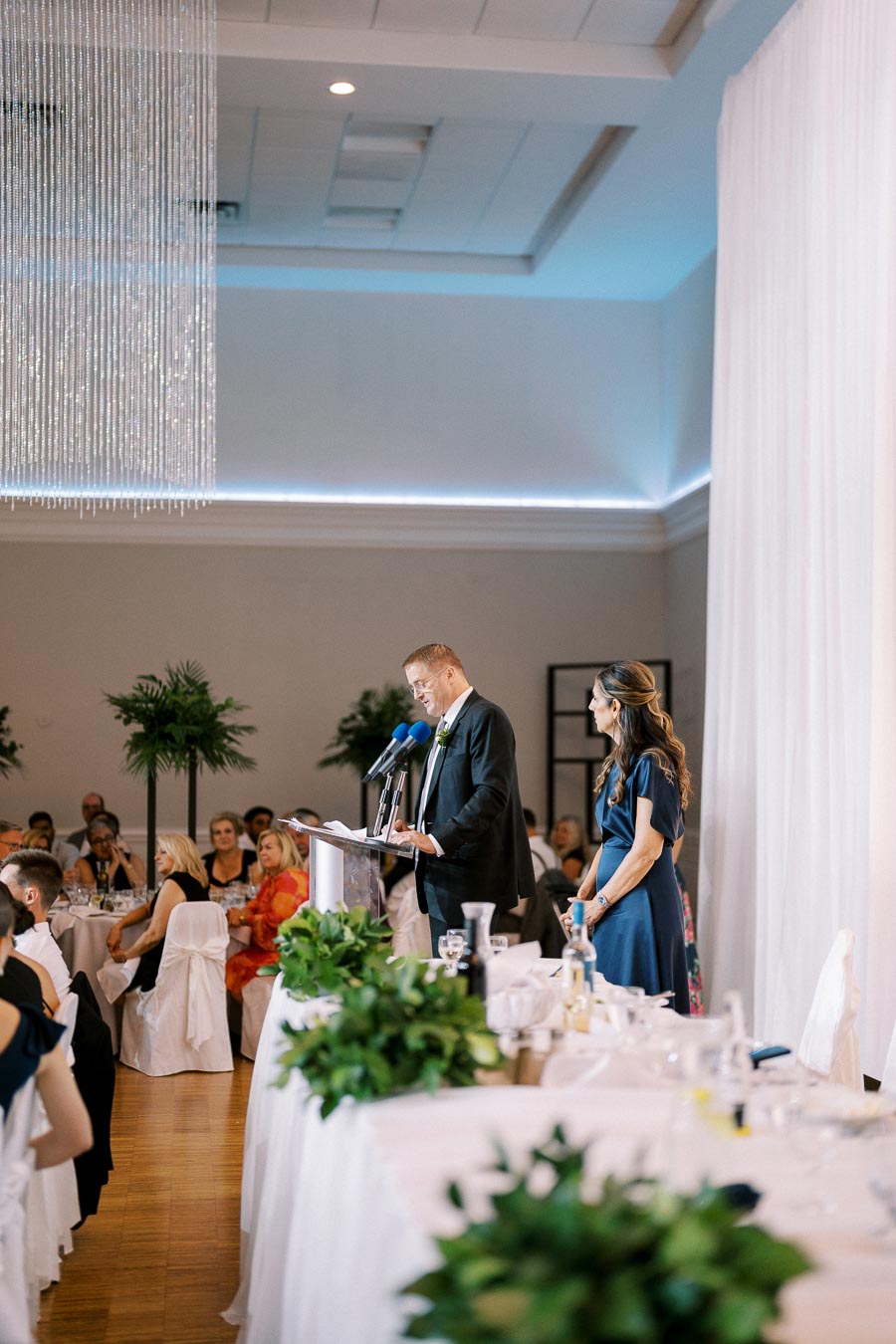 Man and woman speaking at a wedding reception with guests seated at tables, chandeliers and plants in the background.