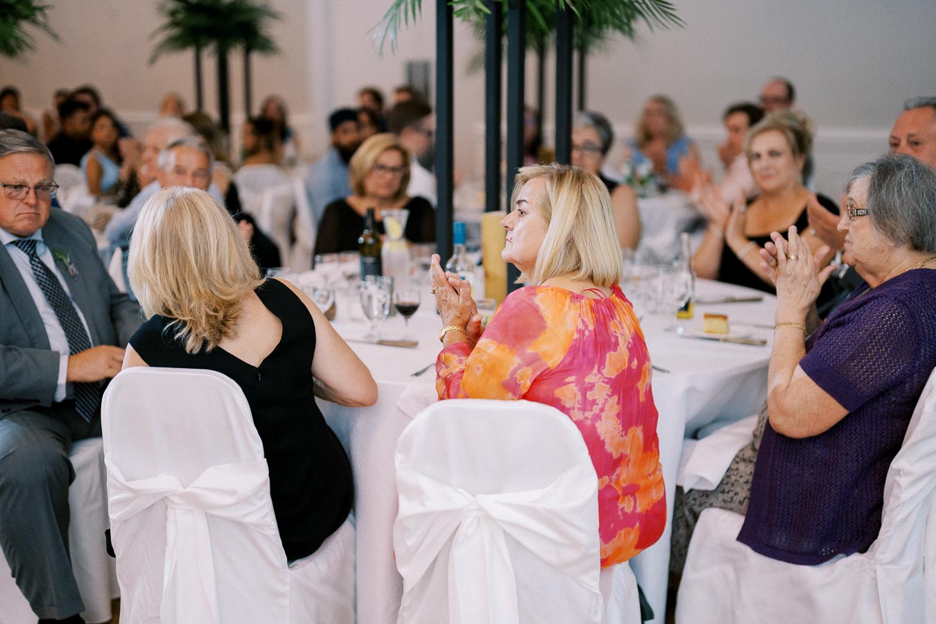 A group of people seated around a decorated table at a formal event, engaging in conversation and clapping, with a backdrop of a lively banquet hall.