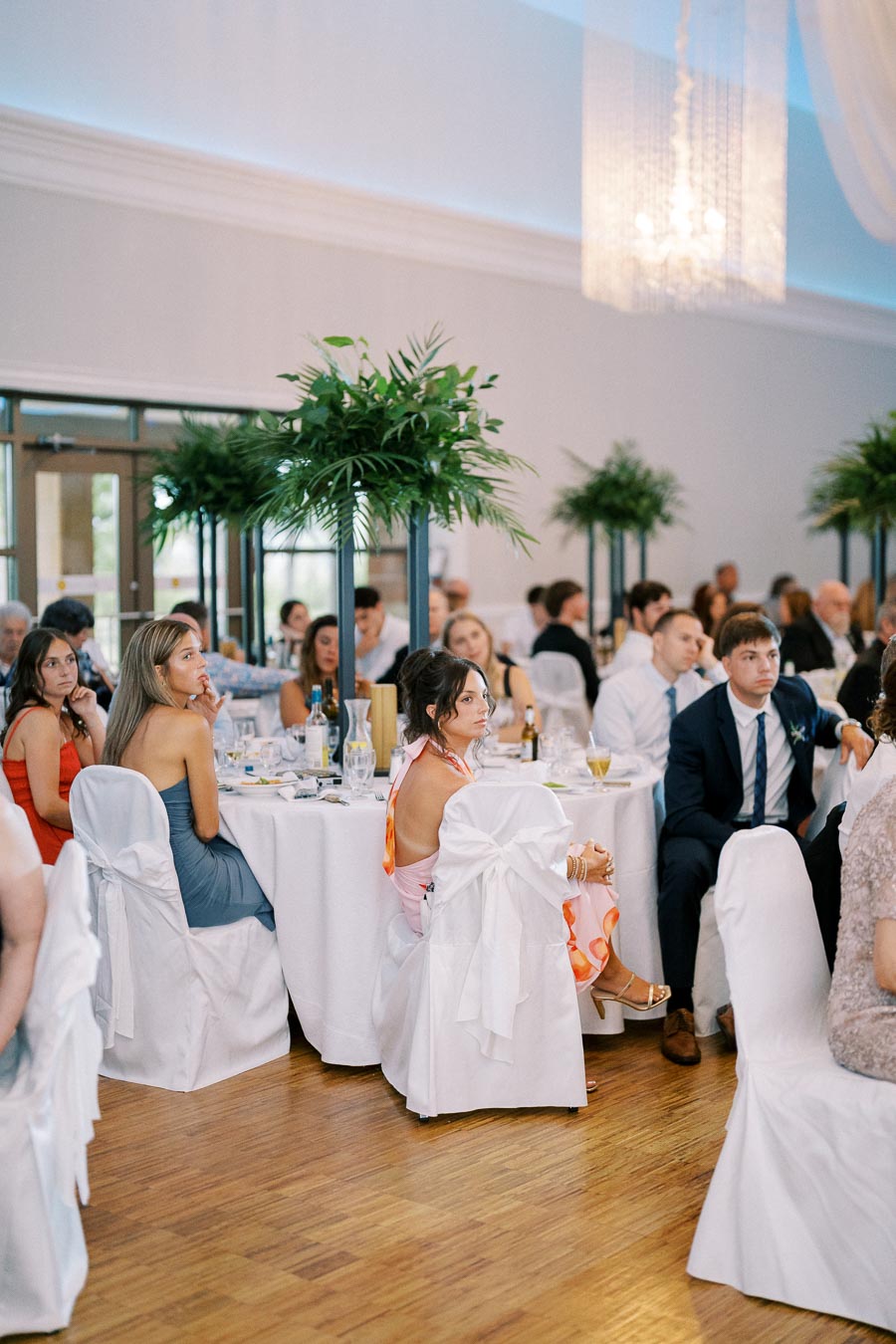 A wedding reception scene with elegantly dressed guests seated at tables adorned with white cloths and decorative greenery centerpieces, in a softly lit venue with a chandelier.