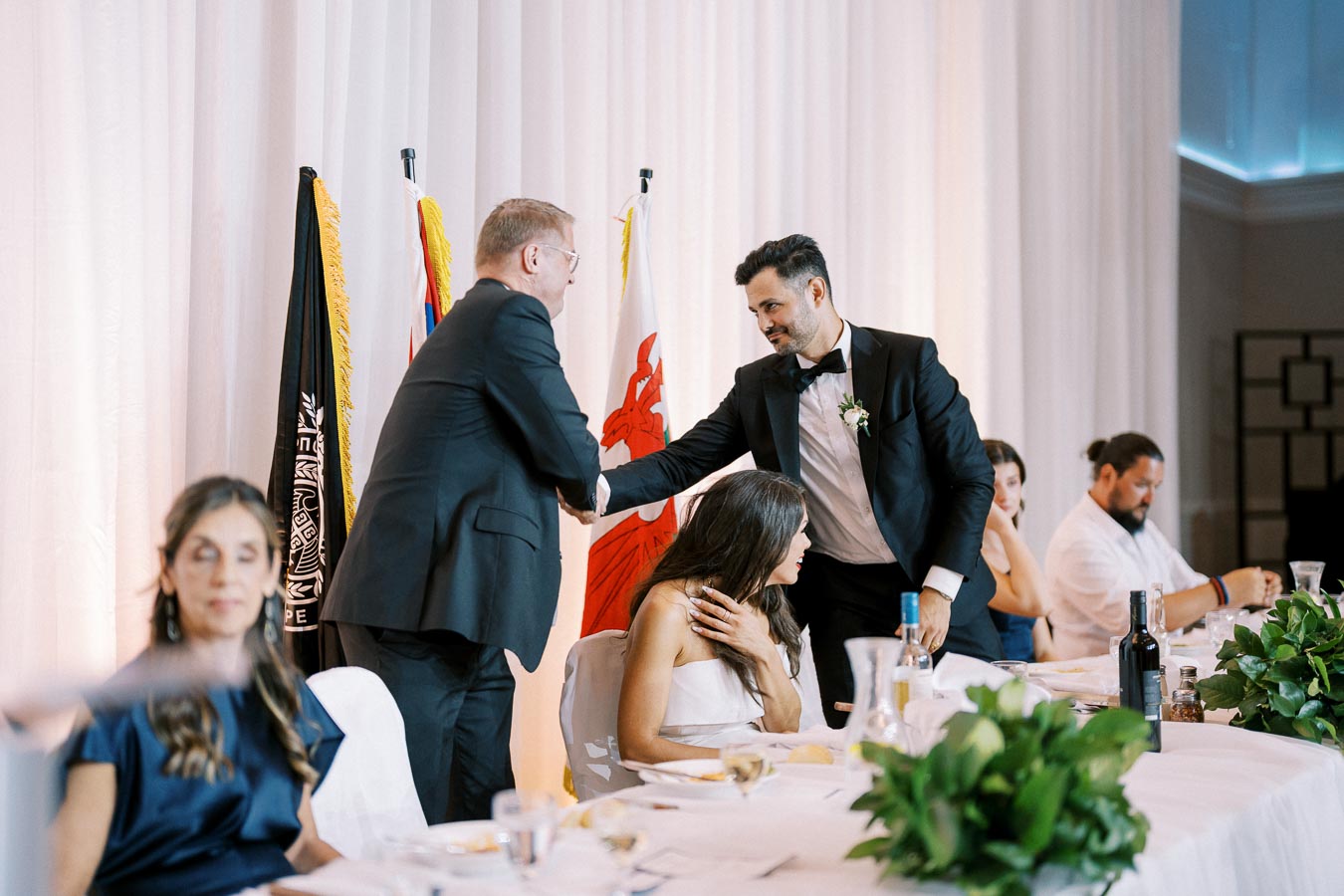 Formal event with two men shaking hands at a banquet table, with elegantly dressed guests seated, featuring national flags in the background and lush table decorations.