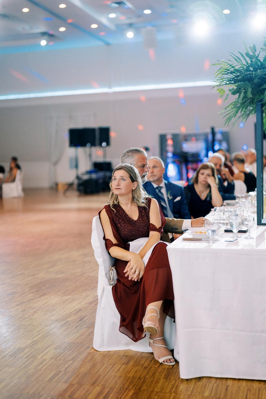 Elegant wedding reception with guests seated at white-covered tables, featuring a woman in a burgundy dress attentively watching an event in a well-lit banquet hall.