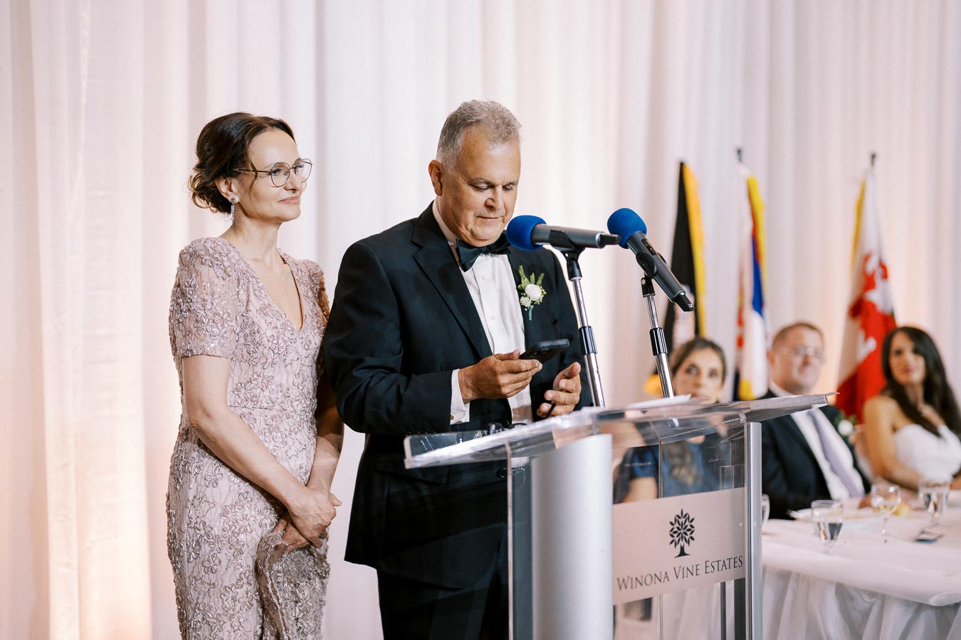 A man in a tuxedo and a woman in an elegant dress stand at a clear podium, delivering a speech at a formal event. The background features a white curtain and various flags. Microphones with blue covers are mounted on the podium.