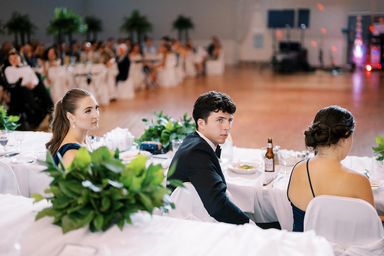 Elegant formal event with attendees seated at a white-clothed table adorned with greenery, focusing on a young woman with a ponytail and a young man in a tuxedo, as an audience in the background listens attentively.