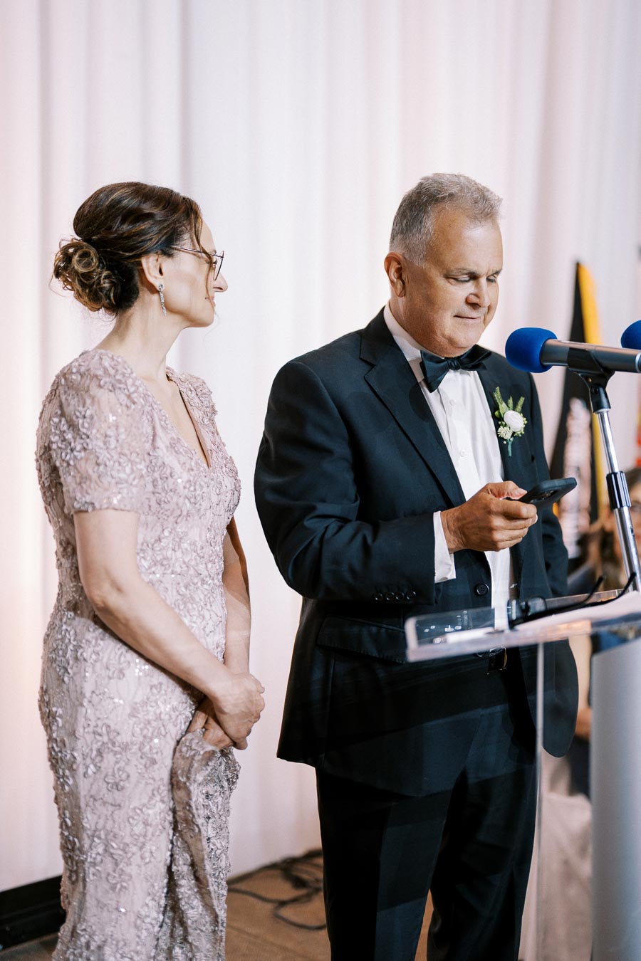 A couple elegantly dressed for a formal event, with the man in a black tuxedo speaking at a microphone while holding a phone, and the woman in a sequined gown standing beside him, in front of a white curtain backdrop.