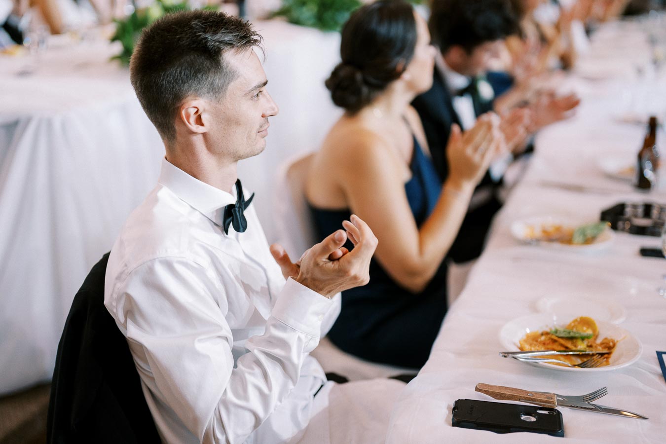 Man in a white dress shirt and bow tie clapping at a formal event, seated at a banquet table with partially eaten plates, surrounded by other guests in elegant attire.