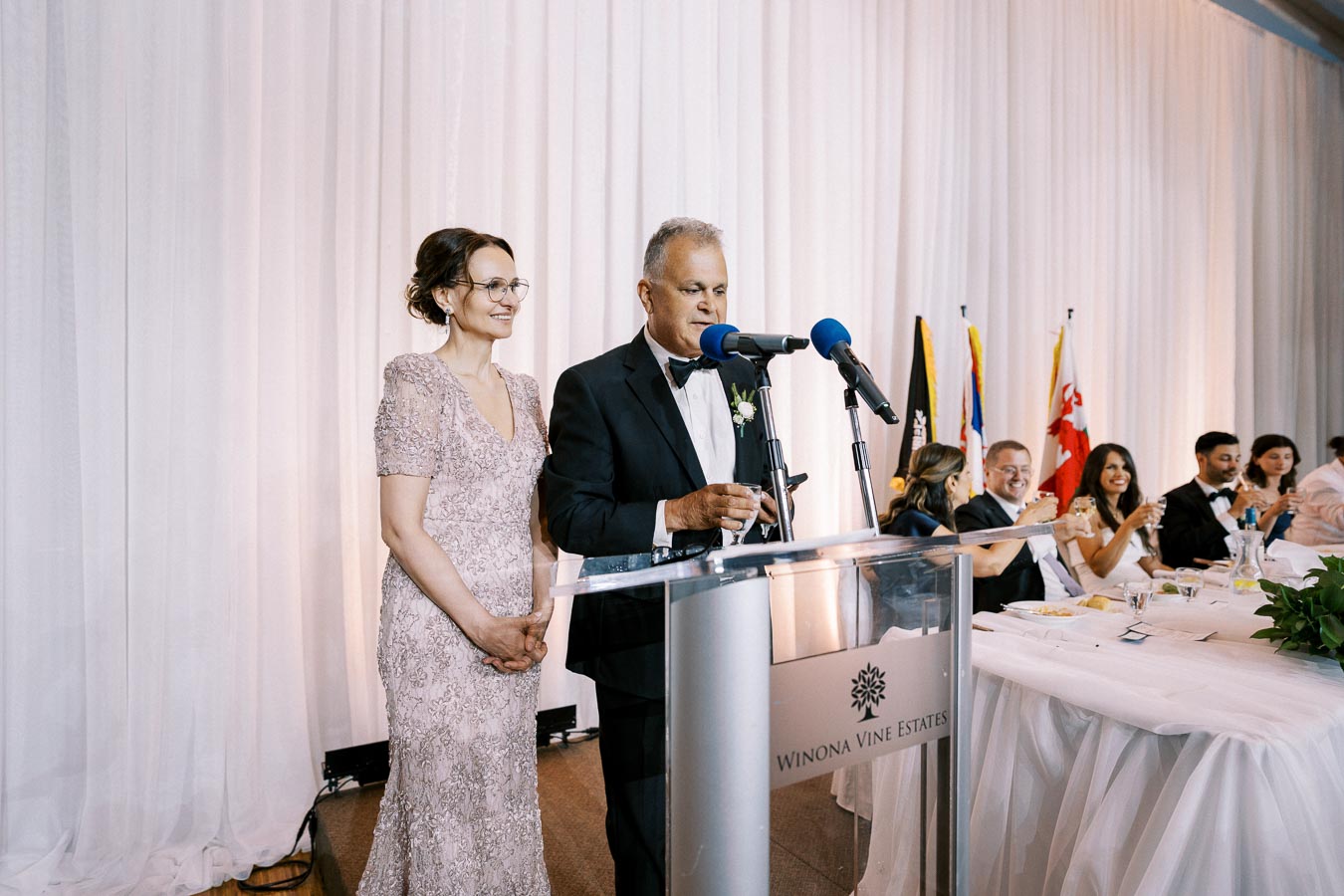 A man and woman stand at a podium delivering a speech at a formal event. The woman is wearing a lace dress, and the man is in a tuxedo. They are in front of a seated audience, who appear to be toasting. Flags are visible in the background. The venue is identified as Winona Vine Estates.