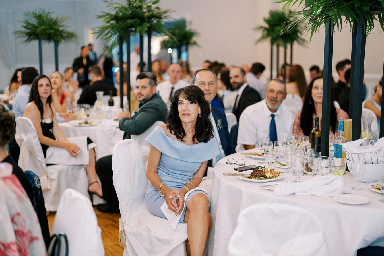 Guests seated at elegantly decorated tables during a formal event, featuring white tablecloths, fine dining settings, and tall green centerpieces.