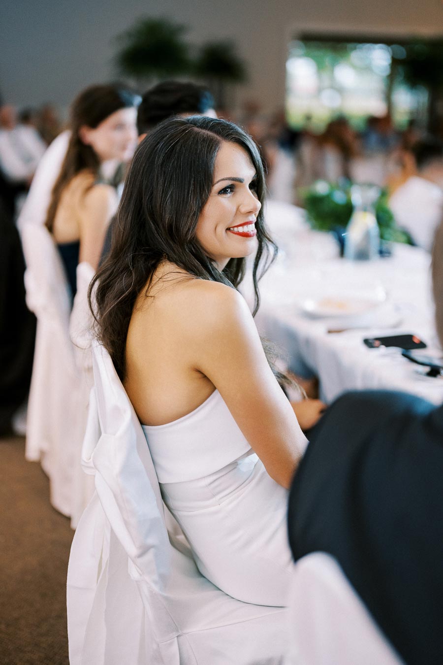 A woman in a white dress smiling at a formal event, seated at a table with a white tablecloth, surrounded by blurred guests in the background.