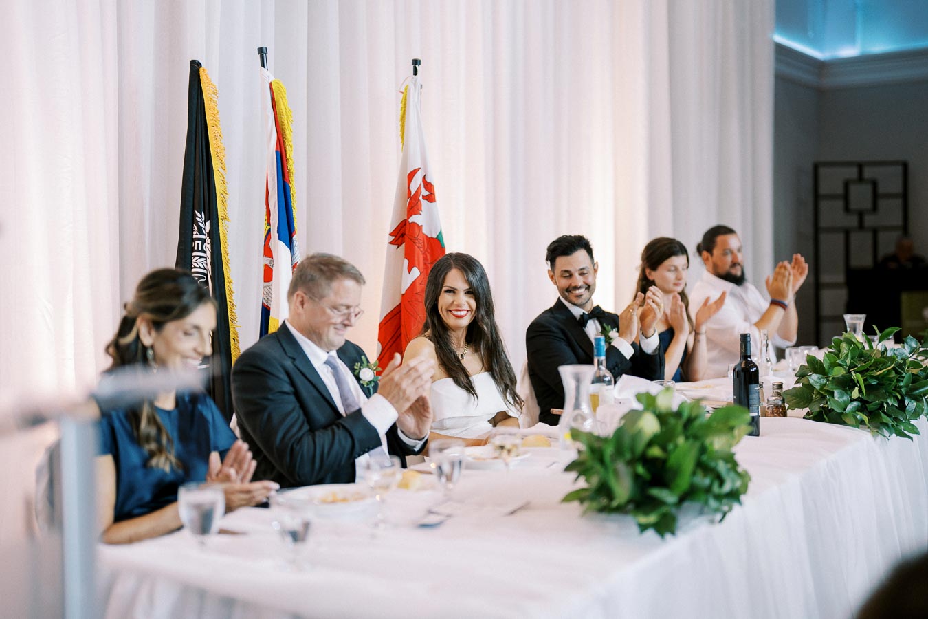 People sitting at a wedding reception table, clapping and smiling, with flags in the background and greenery on the table.