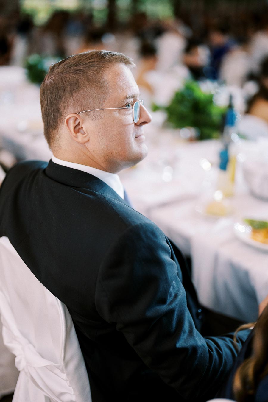 Man in a black suit wearing glasses seated at an elegant banquet table with white tablecloths and floral decorations in a formal setting.