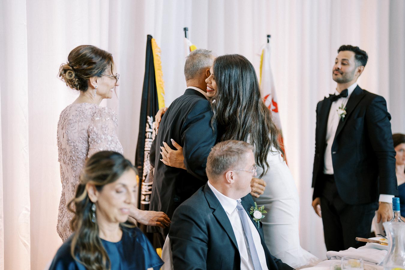 A joyful wedding reception with a bride hugging a guest at a decorated table setup, surrounded by elegantly dressed attendees in a warmly lit venue.