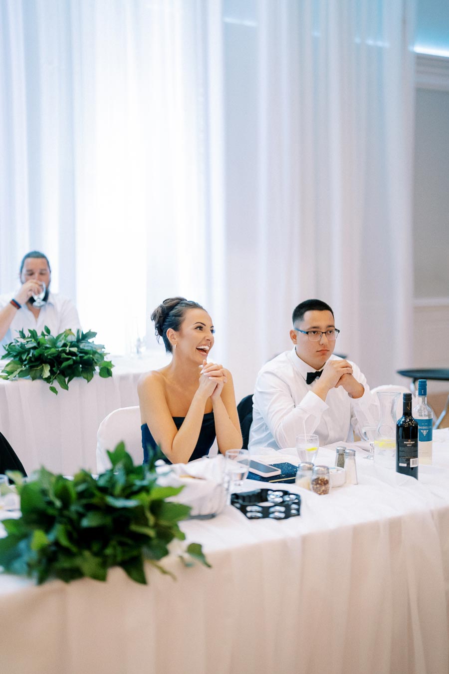 Wedding reception with elegantly dressed guests seated at a table adorned with greenery and wine bottles, engaged in conversation and enjoying the event.