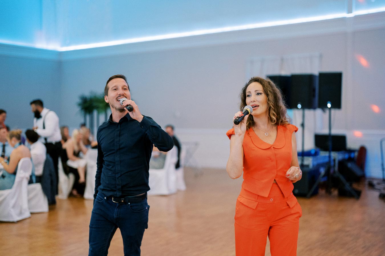 Two singers performing at an indoor event, the woman wearing a bright orange outfit and the man in a dark shirt, with an audience seated at tables in the background.