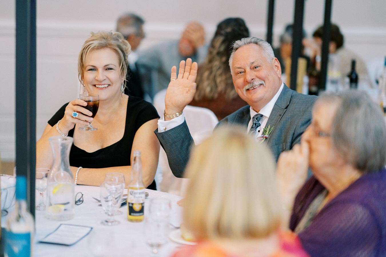 Guests smiling and waving at a table during a joyful wedding reception, with drinks and decorations adding to the celebratory atmosphere.