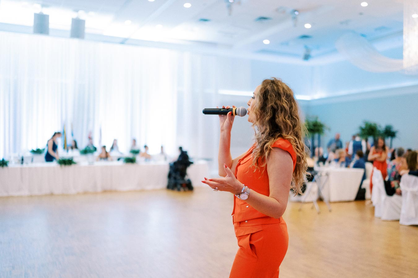 A woman in an orange outfit speaks into a microphone at a formal event in a banquet hall, with an audience seated at tables in the background.