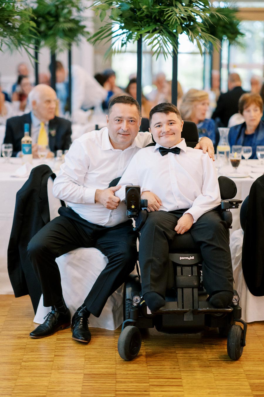 A smiling man in a white shirt poses with a young boy in a wheelchair, both dressed formally, at a festive event with guests and tables in the background.