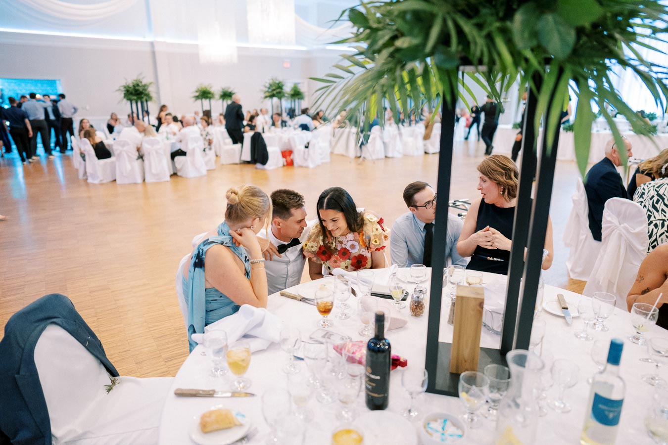 Guests enjoying conversations at a beautifully decorated wedding reception with round tables, elegant white chairs, and lush green centerpieces, creating a vibrant and joyful atmosphere.