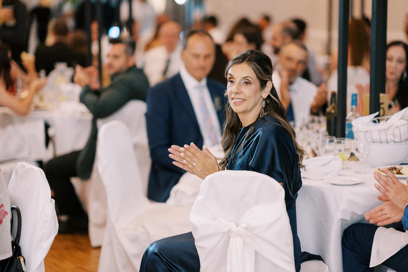 A woman in a navy dress seated at a formal event, clapping her hands surrounded by people at tables covered with white tablecloths and chairs with white covers. The scene conveys a sense of celebration and elegance.