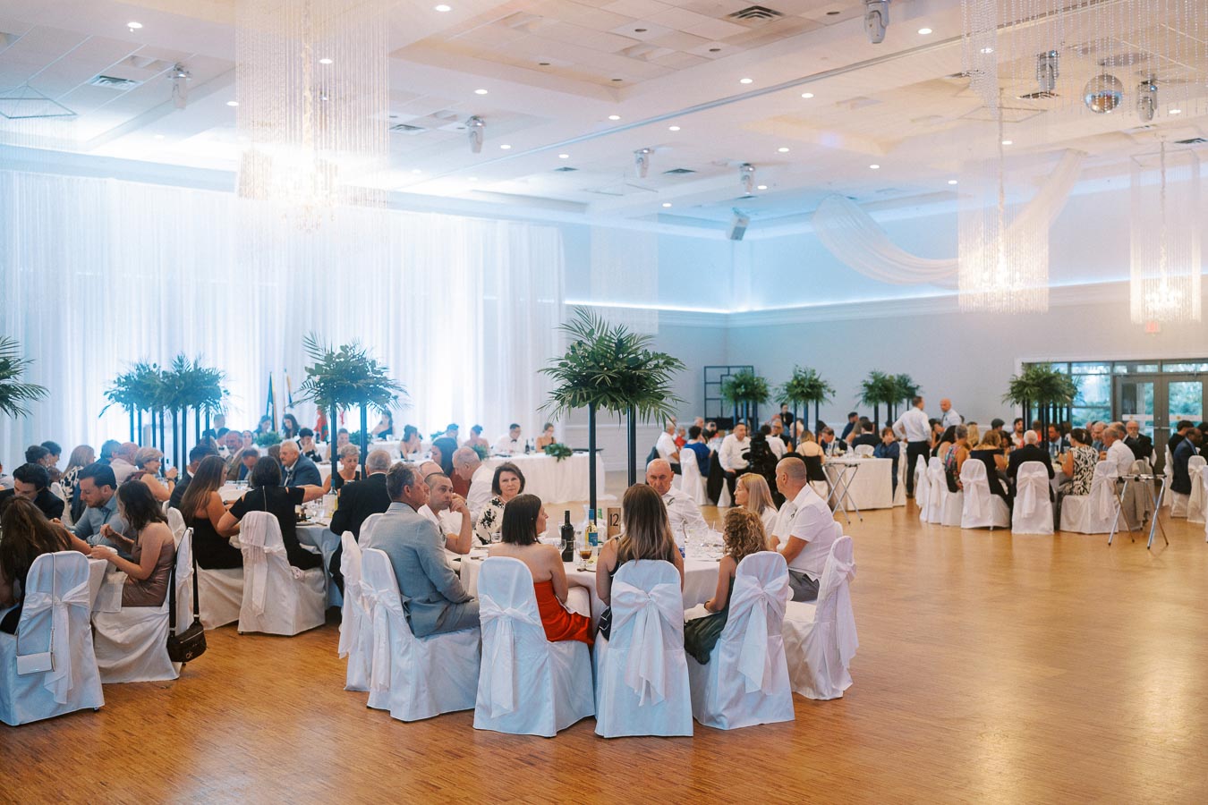 Elegant banquet hall event with attendees seated at round tables, adorned with white chair covers and lush green centerpieces. The room features bright lighting and a modern interior design, creating a sophisticated atmosphere for a formal gathering.