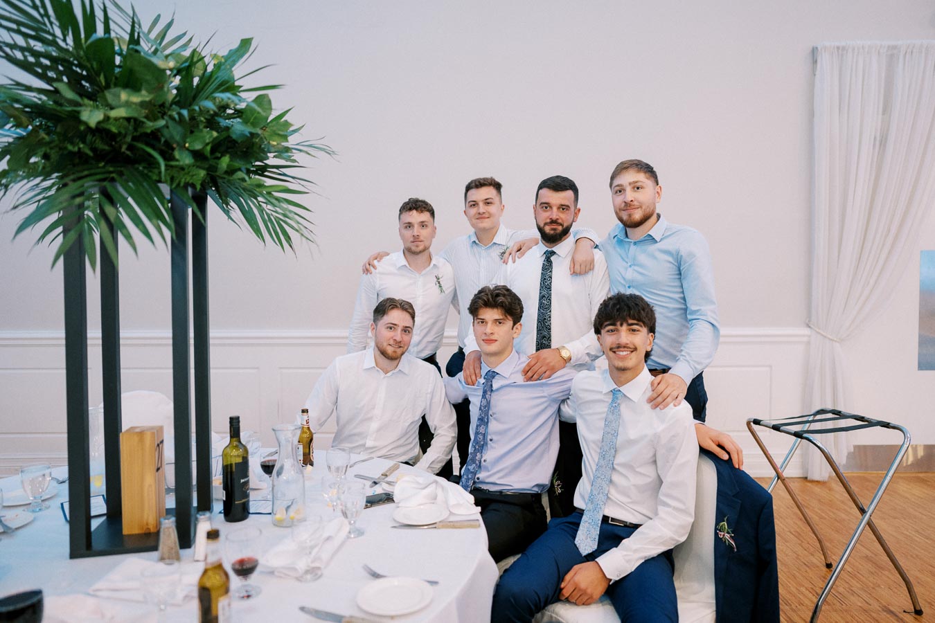 Group of young men dressed in formal attire posing together at a wedding reception, surrounded by dining tables and decorative plants in an elegant banquet hall.
