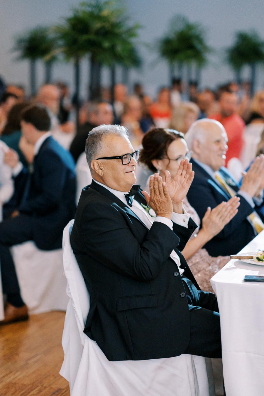 Elderly man in formal attire clapping at a wedding reception, surrounded by other guests seated at tables, with greenery in the background.