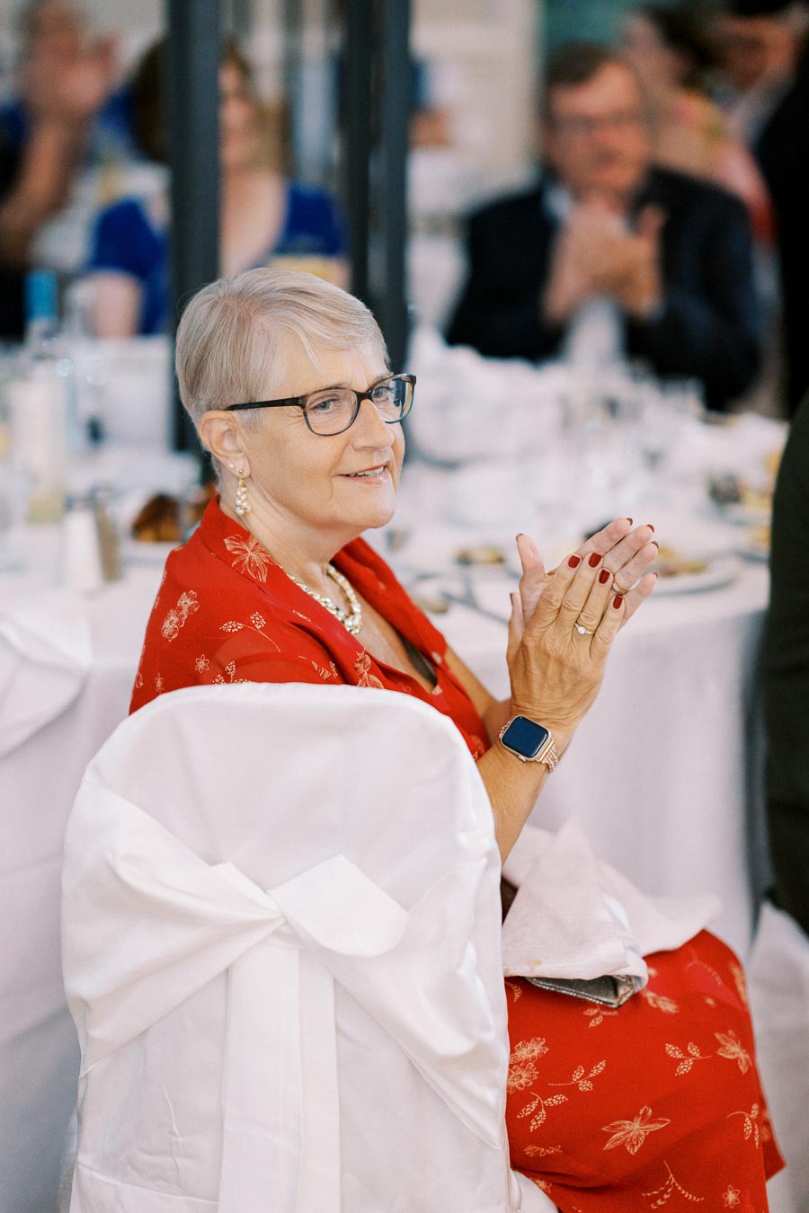Elderly woman with short gray hair and glasses wearing a red floral dress and smart watch, clapping at a formal event with white table settings in the background.