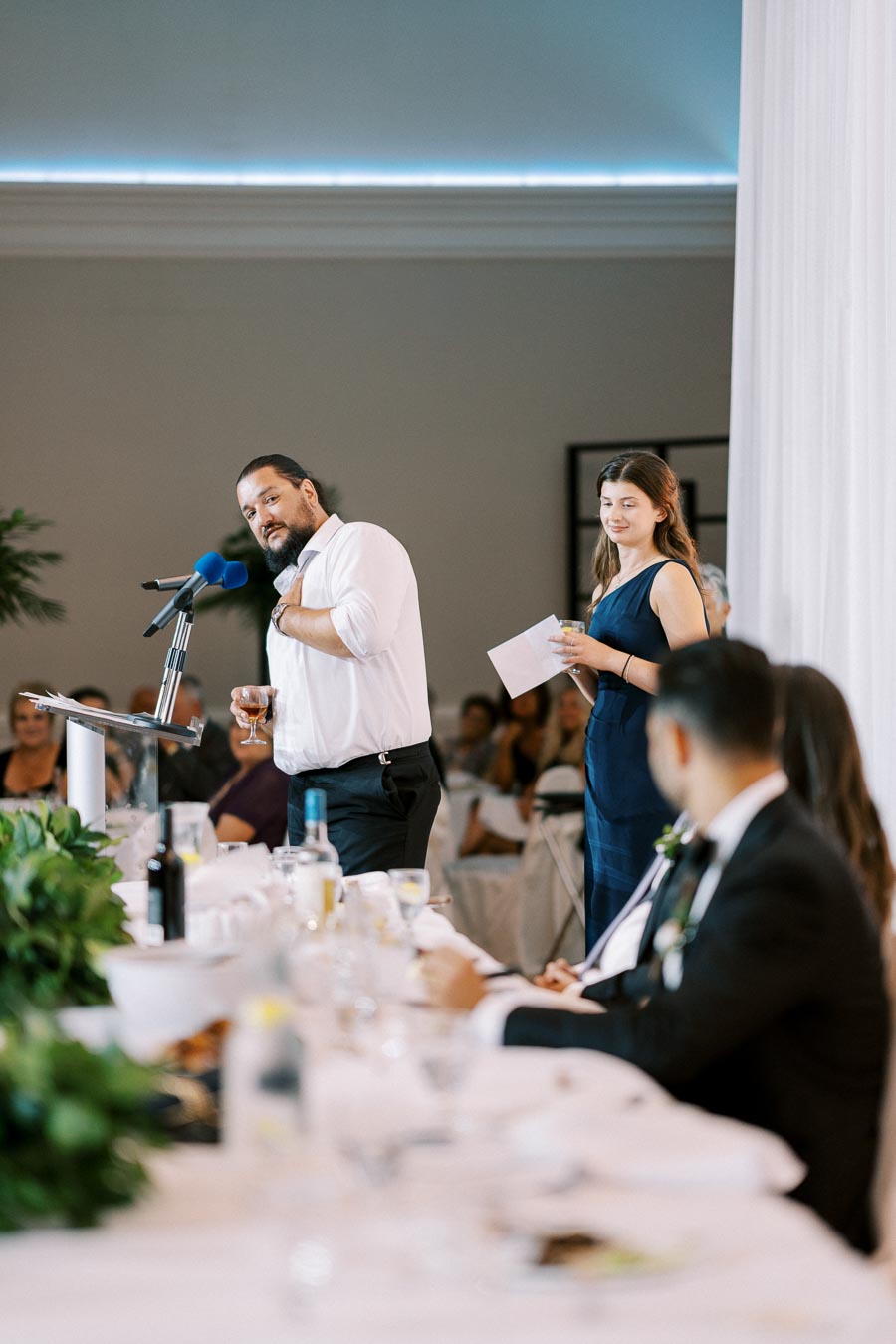 Man giving a speech at a wedding reception, holding a microphone and wine glass, with a woman standing nearby holding a note, in a banquet setting with elegantly set tables in the foreground.