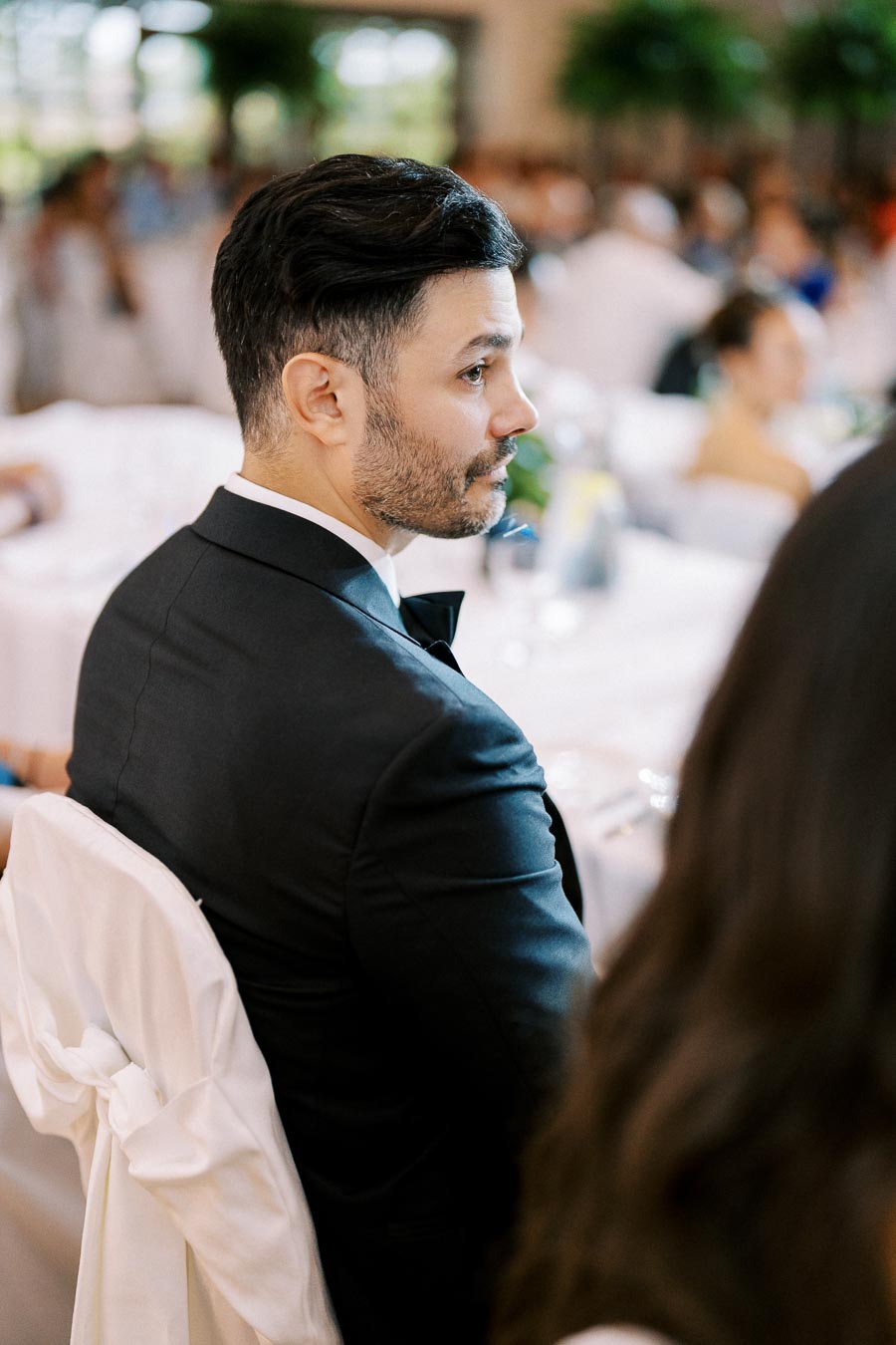 A man in a formal black suit with a bowtie sitting at a decorated table during an indoor event, surrounded by elegantly dressed guests.