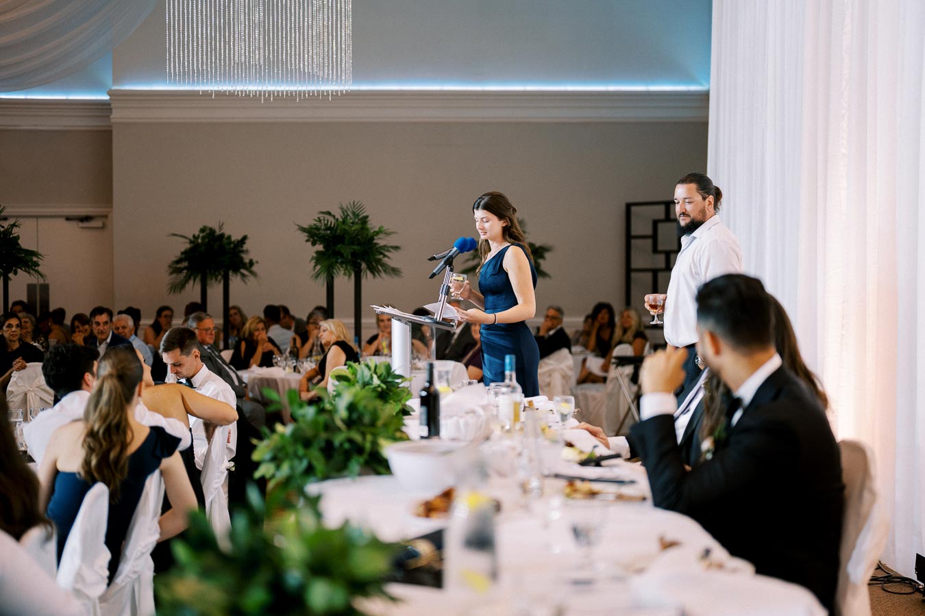 A woman in a blue dress delivers a speech at a formal event, standing at a podium adorned with a microphone. The audience, seated at elegantly set tables with green floral decorations, listens attentively. A man in a white shirt stands nearby, holding a drink. The room features chandeliers and decorative plants.