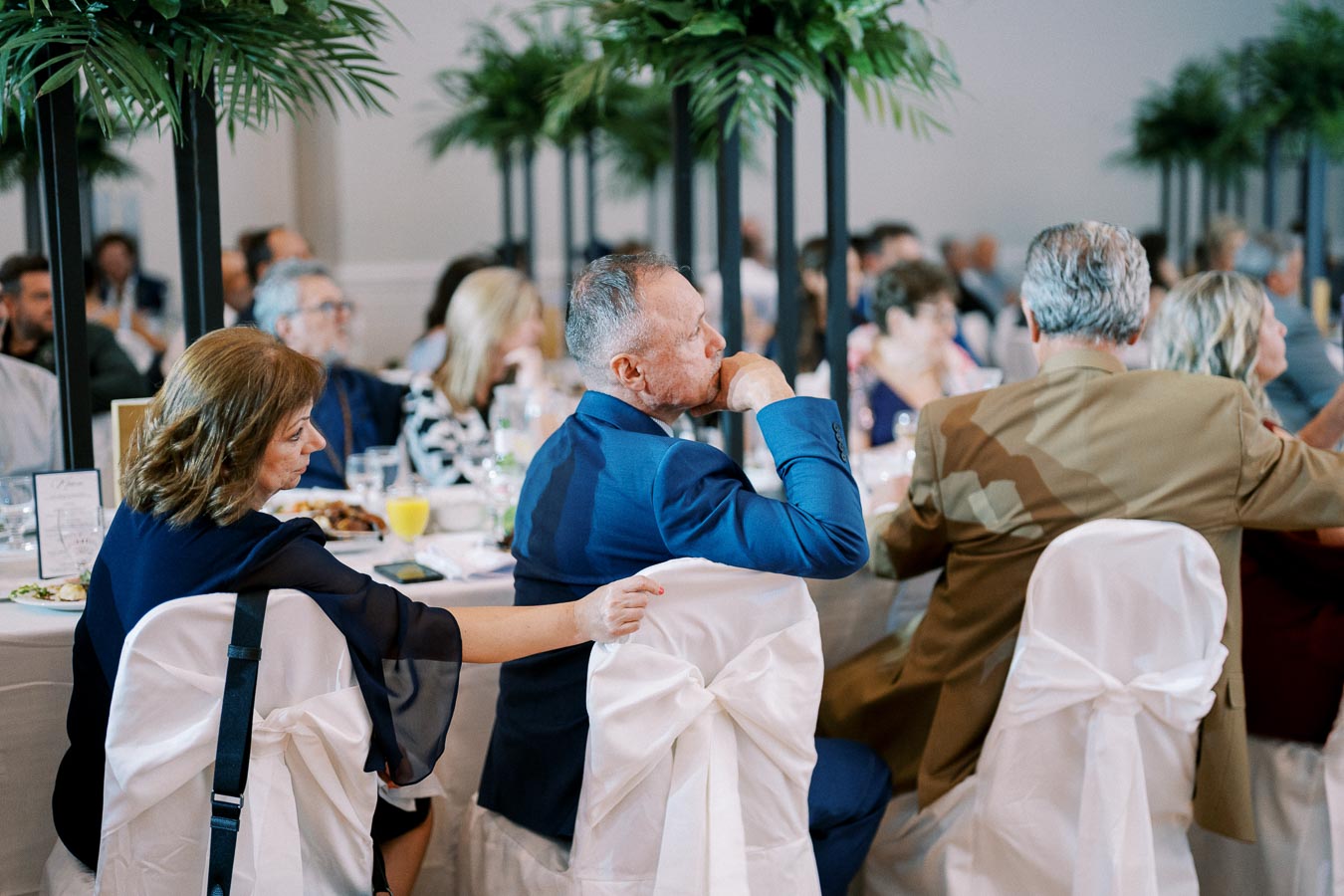 A group of elegantly dressed people attending a formal event, seated at round tables with white tablecloths and greenery decorations.