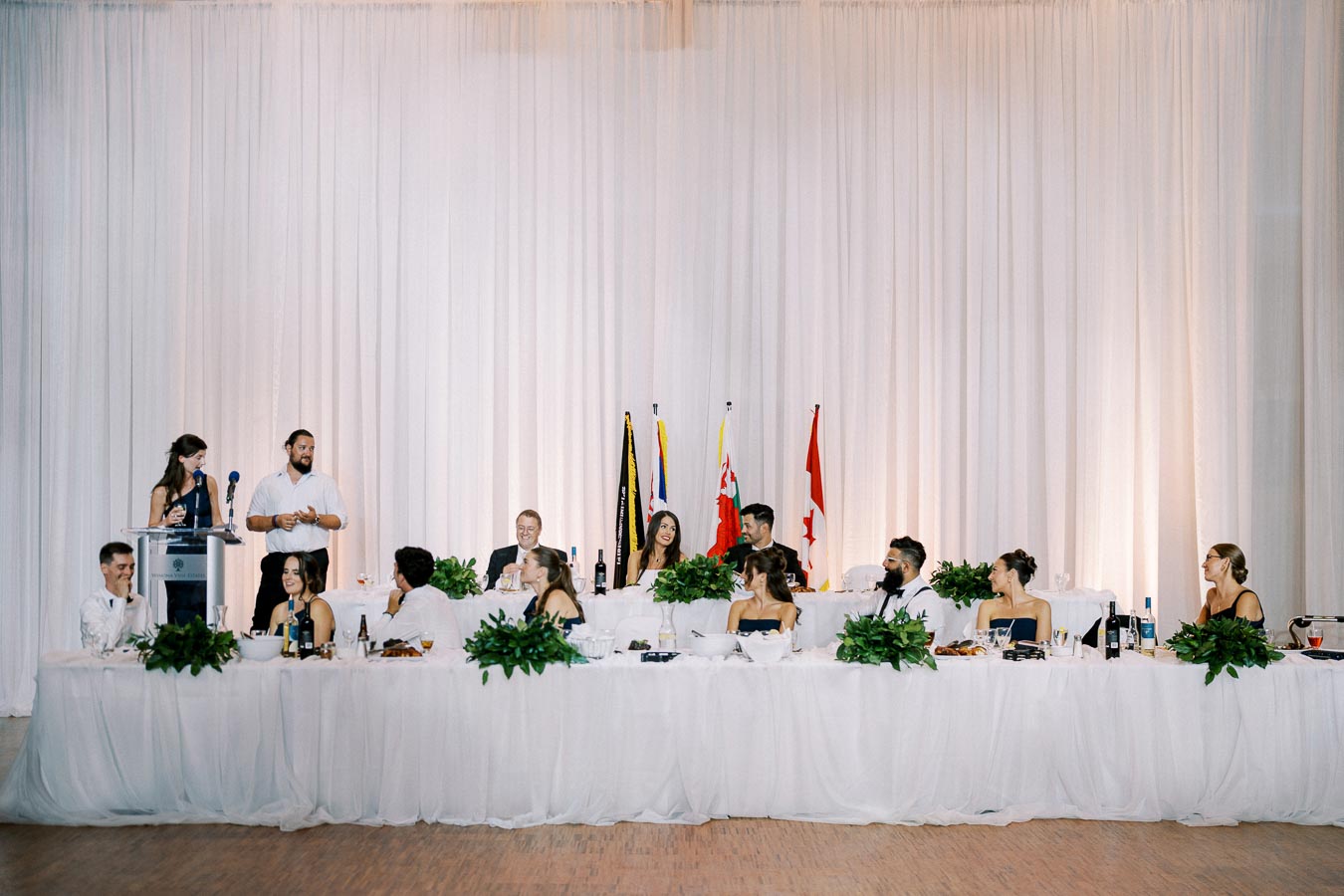 Wedding reception head table with a group of people seated in formal attire. Two speakers stand at a podium on the left, addressing the guests. Several flags are displayed in the background, and greenery decorates the table.