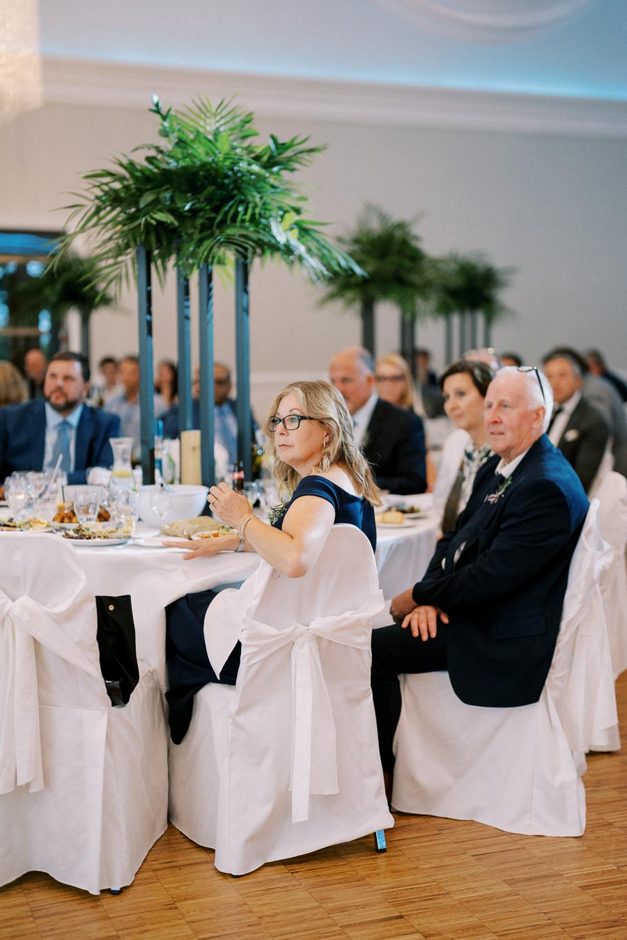 A group of elegantly dressed people seated at a decorated table during a formal event, with lush green centerpieces in a stylish banquet hall setting.