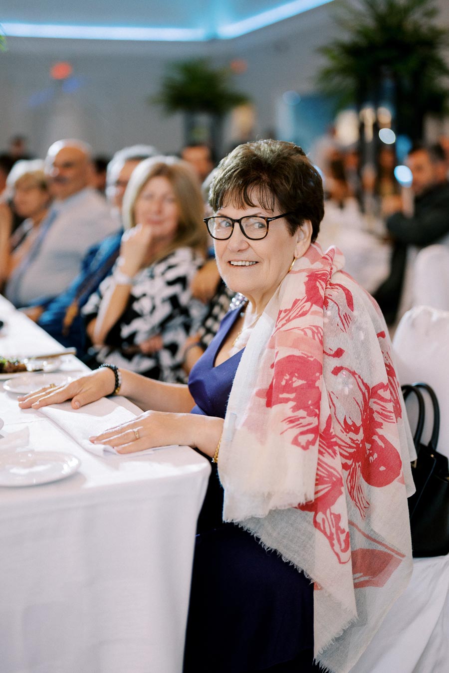 Elderly woman smiling at a formal event, wearing glasses and a floral scarf, seated at a table with other guests in a softly lit banquet hall.