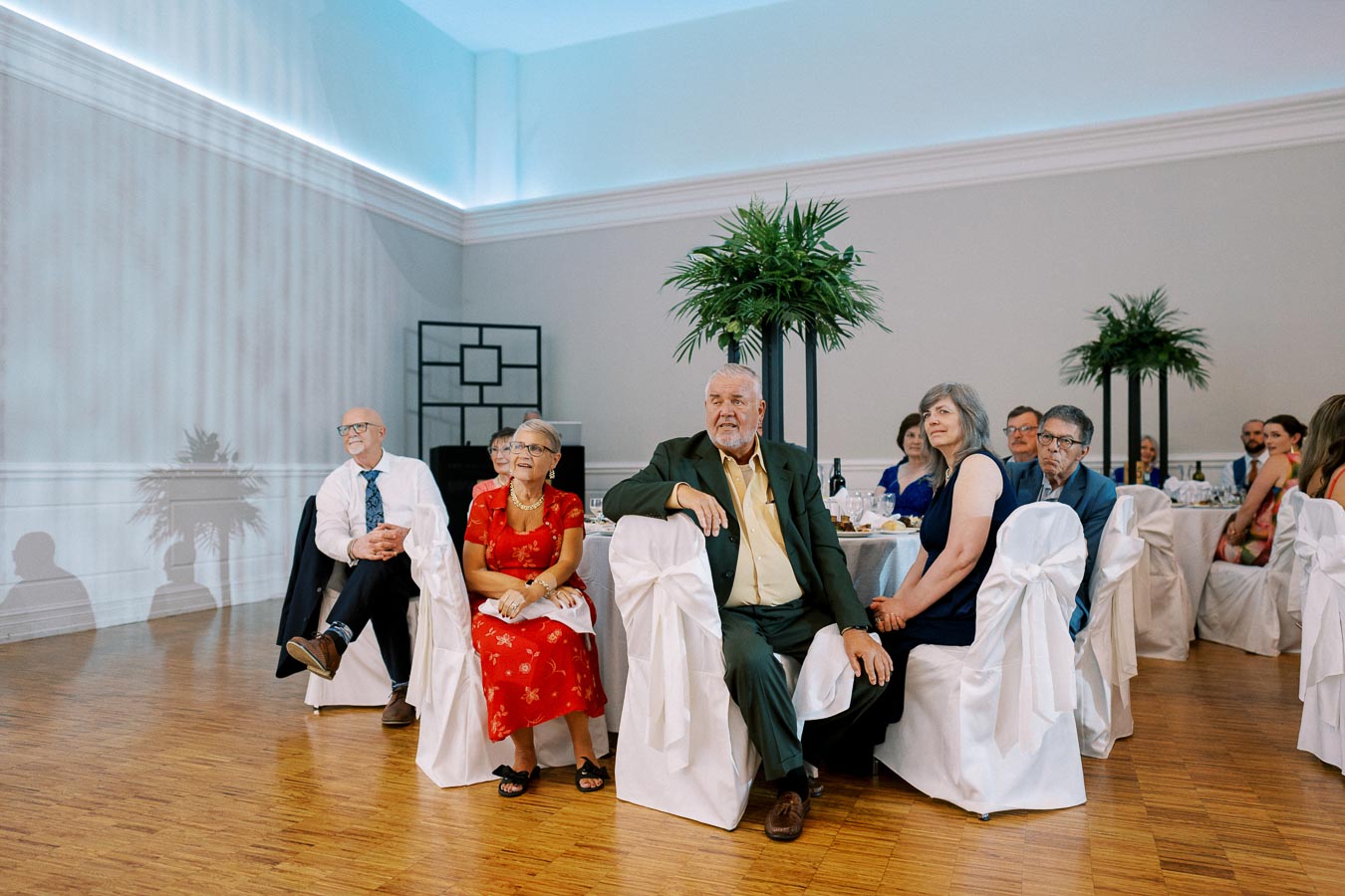 Elderly guests seated at a formal event in a bright, elegantly decorated banquet hall, with white chair covers and decorative plants, attentively watching a presentation.