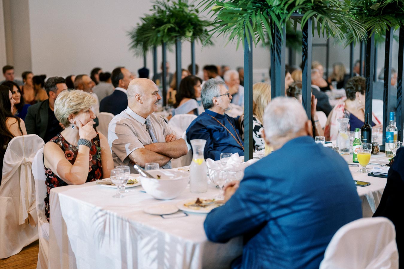 A group of people sitting at elegantly decorated banquet tables during an indoor event, featuring lush green plants as centerpieces and a variety of beverages on the tables.