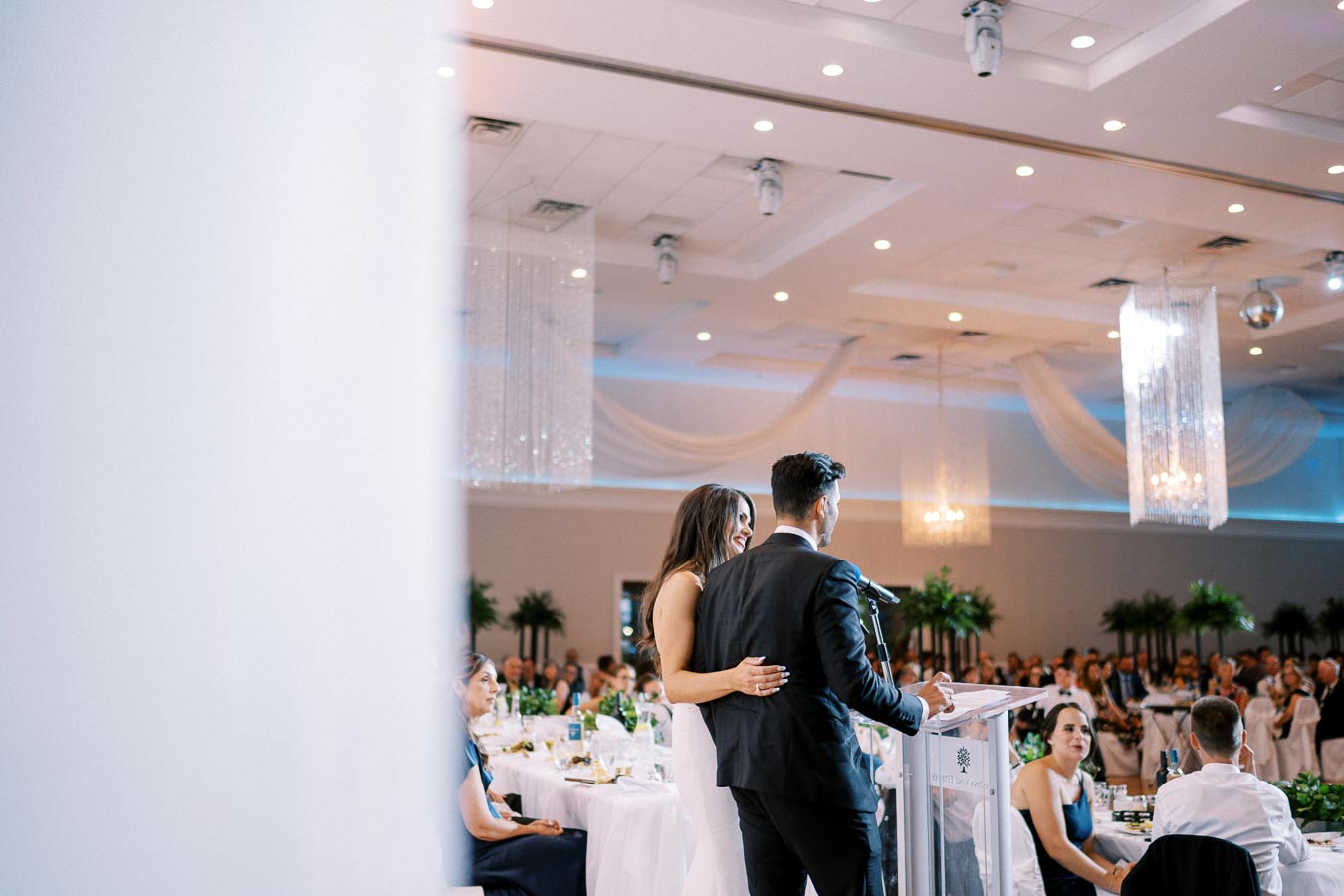 Couple giving a speech at a wedding reception in a decorated banquet hall, with guests seated at elegantly set tables.