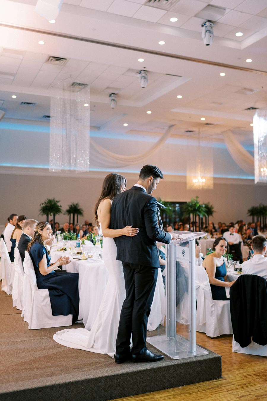 Wedding reception with a bride and groom giving a speech at the podium, surrounded by guests seated at elegantly decorated tables in a banquet hall.