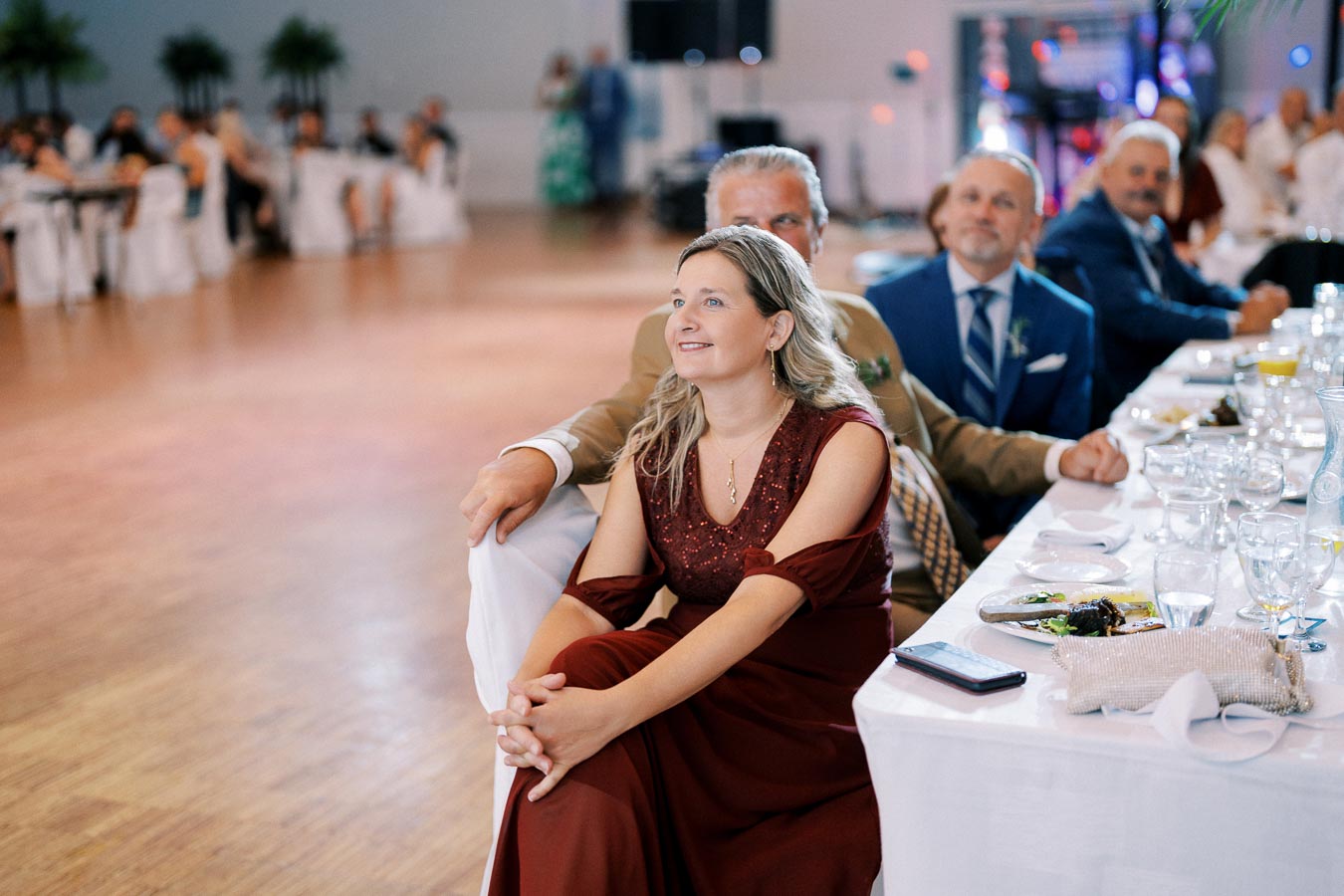 A woman in a burgundy dress sits smiling at a wedding reception table, surrounded by elegantly dressed guests in a decorated hall.