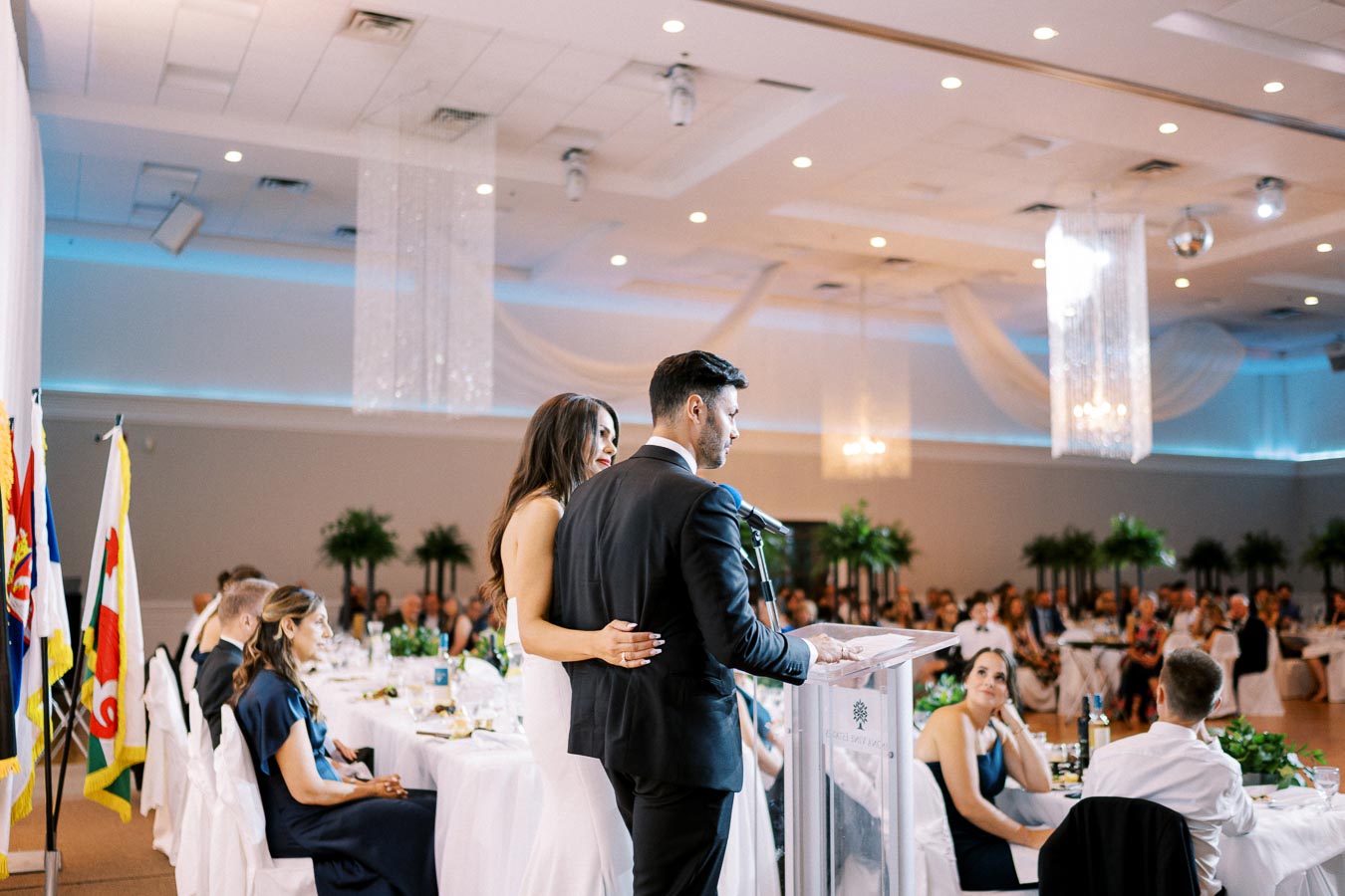 A couple giving a speech at a wedding reception in a decorated banquet hall, with elegantly dressed guests seated at tables and flags displayed in the background.