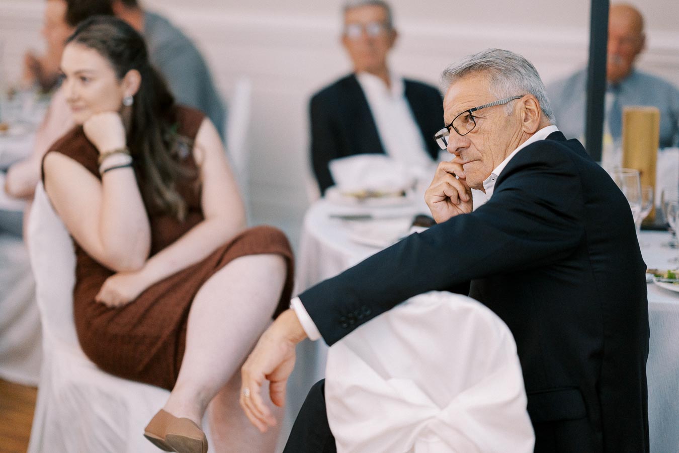 Senior man in a suit attentively listening during a formal event, surrounded by other seated guests.