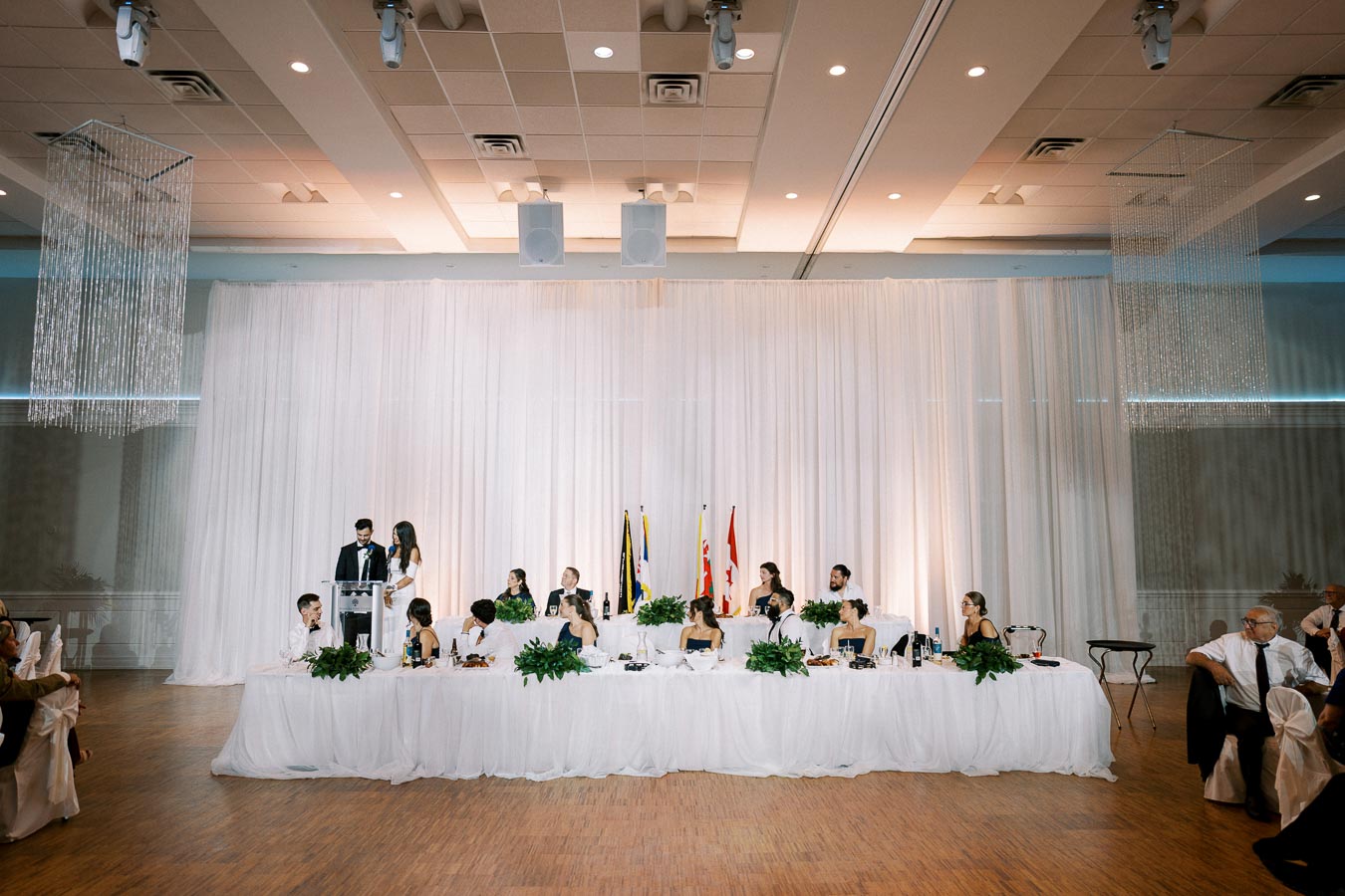 Wedding reception hall with a long head table covered in white cloth, decorated with greenery; a couple is standing at a podium, surrounded by elegantly dressed guests seated, with flags visible in the background.