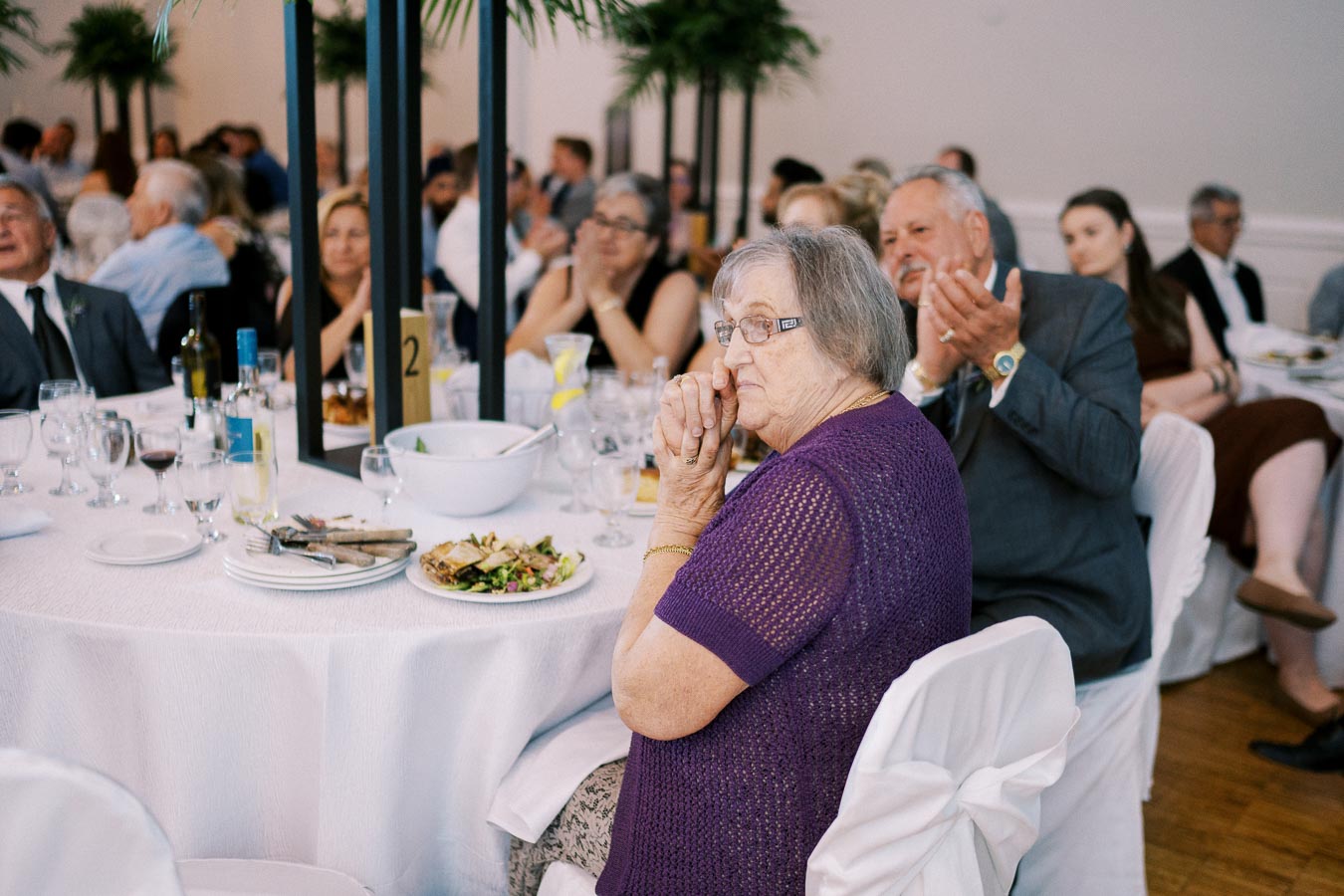 A group of elegantly dressed people sitting around a table at a formal event, with an elderly woman in a purple sweater in the foreground, surrounded by plates, glasses, and a white tablecloth, emphasizing a celebratory gathering.