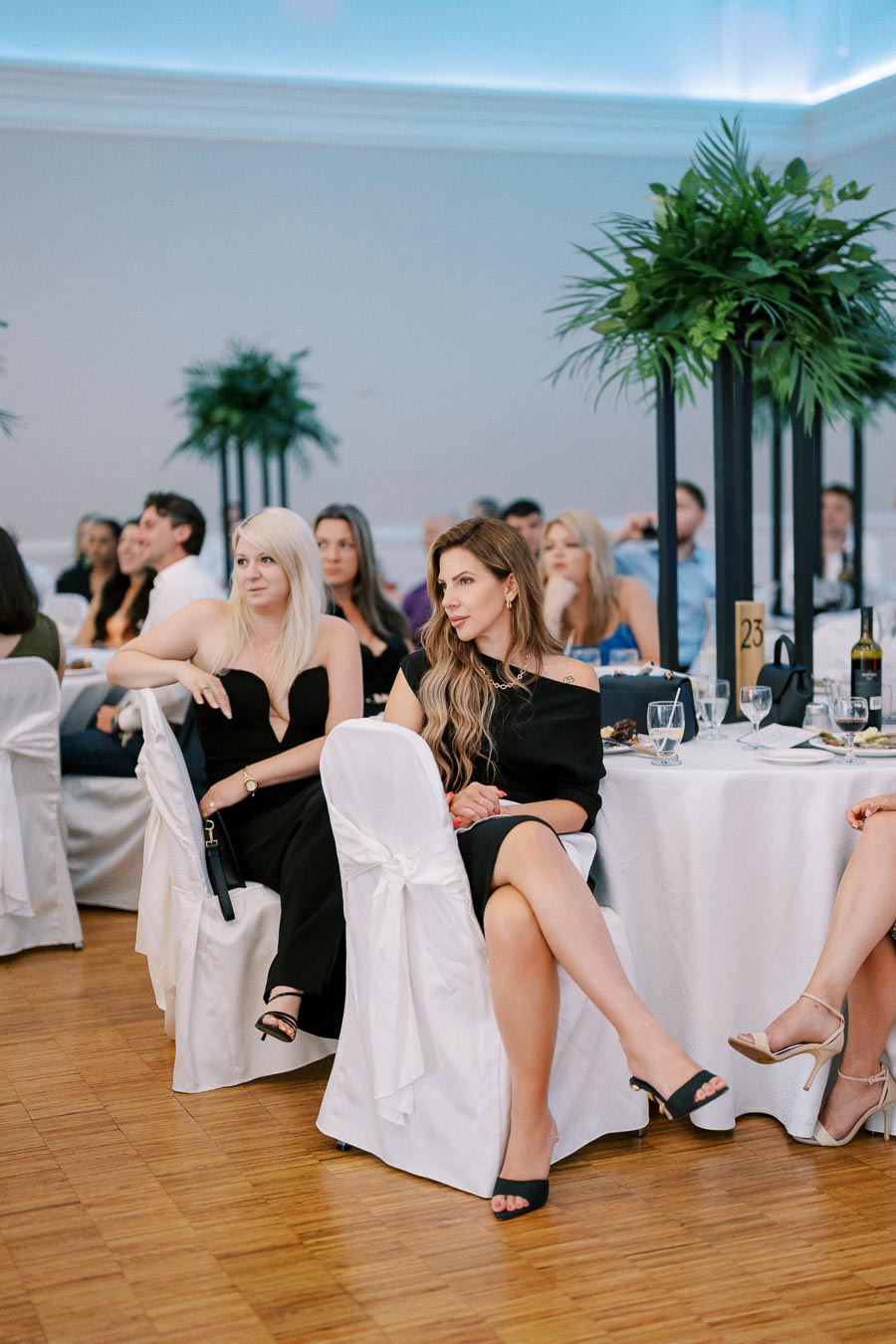 A group of elegantly dressed people seated at a formal event, wearing black dresses, with tables covered in white tablecloths and decorated with tall green plants in the background.