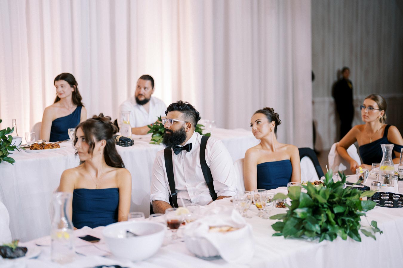 Wedding reception with guests seated at elegantly decorated tables, featuring navy blue attire and lush green centerpieces.