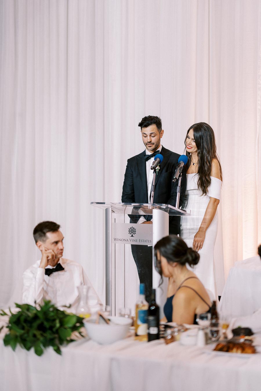 Wedding reception at Winona Vine Estates featuring a couple giving a speech at the podium, with elegantly dressed guests seated at tables adorned with greenery and wine.