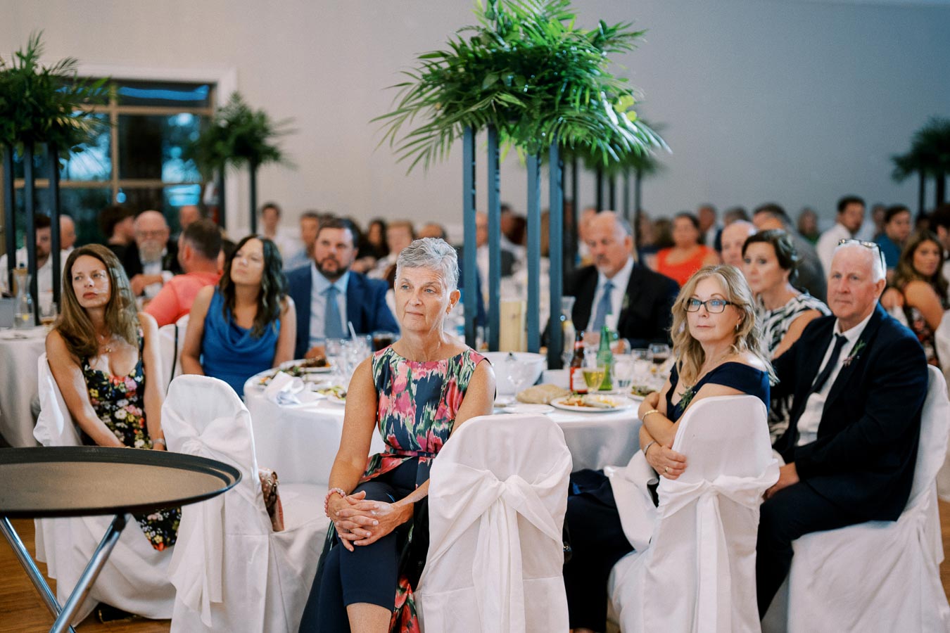 Guests attentively listening during an elegant indoor event with round tables, vibrant floral centerpieces, and formal attire.