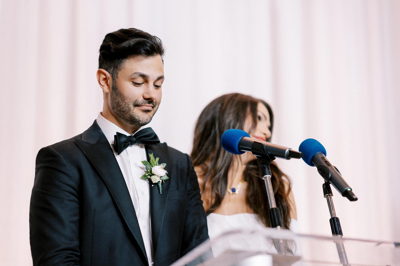 A groom in a black tuxedo and bow tie, standing next to the bride at a wedding ceremony, speaking into a microphone with a backdrop of white drapes.